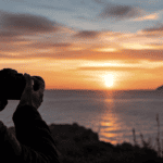 Mani che tengono una macchina fotografica abbassata davanti a un paesaggio di borgo italiano al tramonto